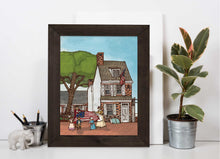 Framed artwork of  the Betsy Ross house in Philadelphia with people and American flags on a white surface with a plant and small elephant figurine.